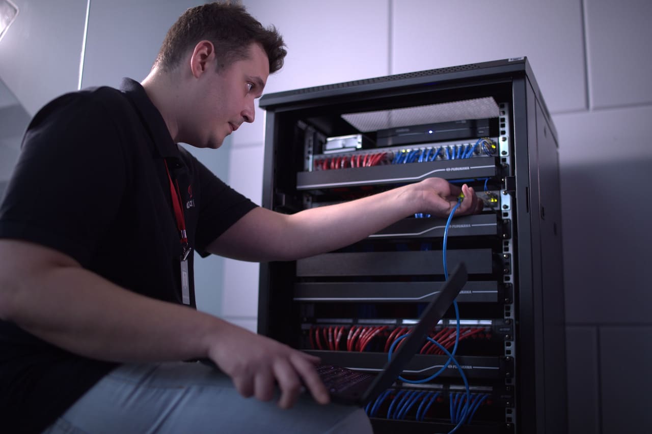 Técnico da AtentoTI com notebook no colo, conectando um cabo no rack de rede do cliente.