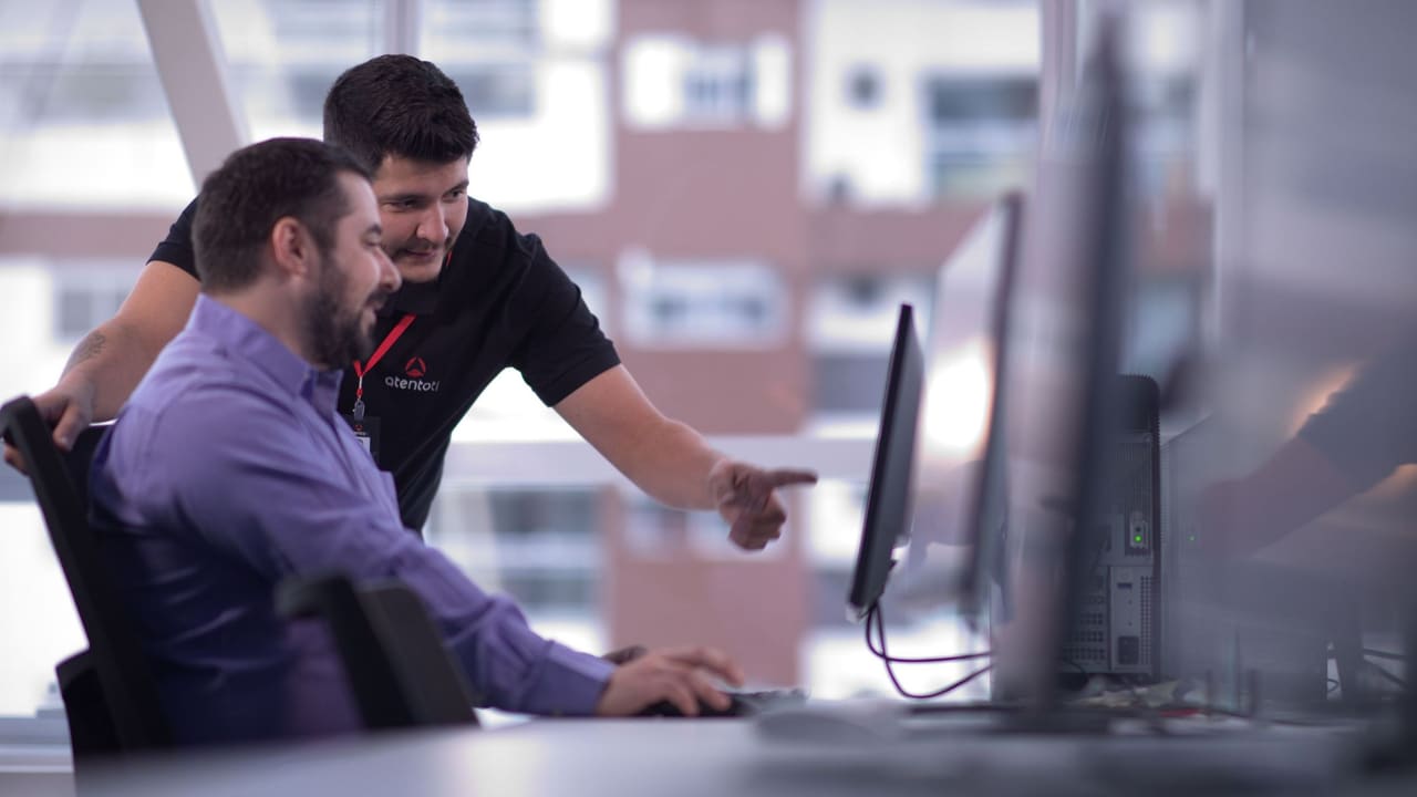 Técnico da AtentoTI ao lado de uma pessoa na mesa dela, apontando para a tela do computador.